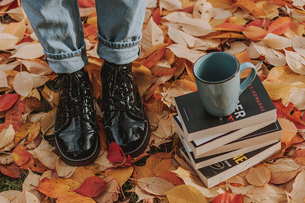 Częściowy widok na osobę stojącą w jesiennych liściach. Obok leżą książki i stoi kubek. fot Pexels Rende